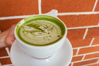A hand presents a beautiful matcha latte with delicate foam art, set against a charming brick wall backdrop.