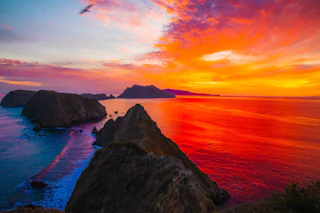 Stunning rocky coastline at sunset, with brilliant orange, red, and purple sky mirrored on the calm ocean waters.