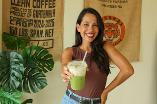 Happy woman enjoys a refreshing iced green drink, surrounded by coffee sacks and lush greenery, creating a lively scene.