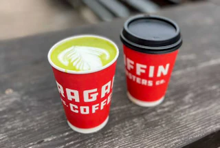 Two vibrant red coffee cups on a wooden bench, one with beautiful latte art and the other ready to go with a lid.