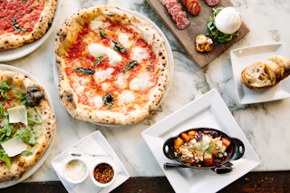A delicious spread of two gourmet pizzas, a cheese plate, roasted vegetables, bread, and condiments on an elegant marble table.