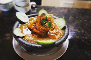 Delicious bowl of shrimp in flavorful sauce, beautifully garnished with cilantro and cucumbers, served on a white plate.