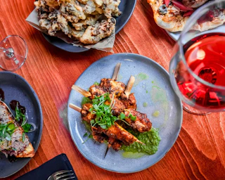 Inviting plate of skewered grilled meat with green sauce, wine, fresh bread, and delicious appetizers on a rustic wooden table.