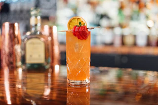 A vibrant orange cocktail topped with a dried lime slice and fresh berries sits on a wooden bar, creating an inviting atmosphere.