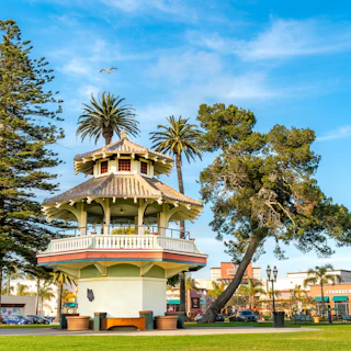A charming white gazebo in a lush park with palm trees, nearby shops, and Starbucks under a bright blue sky.