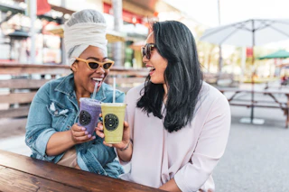 Two women enjoy a sunny day outdoors, smiling and sipping smoothies together at a wooden table in a welcoming setting.
