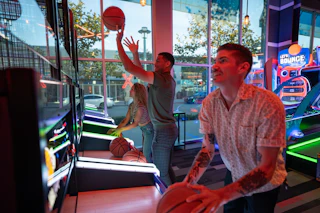 Visitors enjoying arcade basketball games together under vibrant neon lights at a lively entertainment venue.