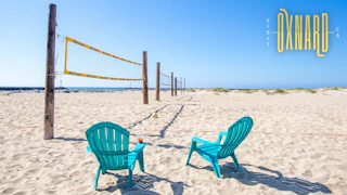 Two turquoise chairs on sunlit sand face a volleyball net under a clear sky; "Visit Oxnard, CA" welcomes visitors.