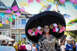 A vibrant Day of the Dead celebration features a person in ornate costume and makeup outdoors, with festive banners and joyful crowds.