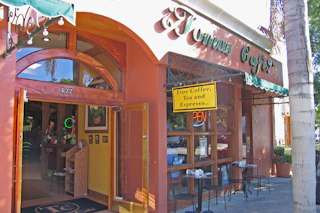 Charming cafe exterior with welcoming arched doorway, vibrant green awning, outdoor tables, and a sign for premium drinks.
