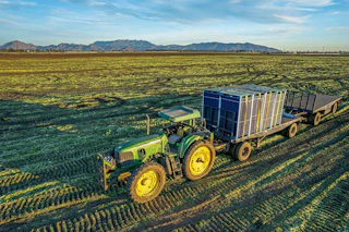 A vibrant green tractor pulls trailers with crates across a scenic field, framed by majestic mountains and a clear blue sky.
