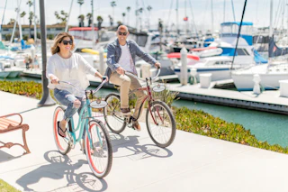 Two people happily ride bikes along a sunny marina with boats and palm trees in the background, enjoying a perfect day.