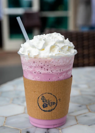 A refreshing pink iced drink with whipped cream in a plastic cup and brown sleeve, beautifully presented on a marble table.