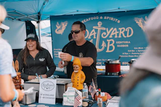 Smiling staff serve delicious chowder at a welcoming Boatyard Pub booth, decorated with nautical accents and American flags.