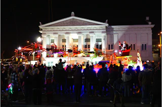 Santa waves joyfully from a sparkling sleigh float with reindeer at a festive night parade, crowds cheering near a grand landmark.