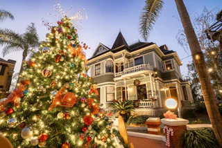 A beautifully lit Christmas tree with vibrant ornaments stands before a festive, decorated Victorian house at dusk, radiating holiday cheer.