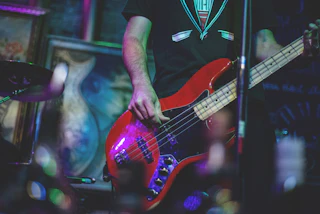 A musician in a black shirt plays a red electric bass guitar on stage, surrounded by vibrant lights and artistic backdrops.