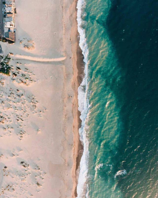 Aerial view of pristine waves gently washing onto a golden sandy beach, with lush greenery and inviting buildings on the left side.