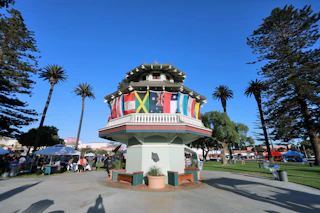 A vibrant pavilion adorned with international flags welcomes visitors to a sunny, palm-lined park bustling with happy people.