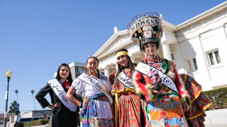 Four women in colorful traditional Mexican dresses and sashes smiling outside a charming historic building on a beautiful sunny day.