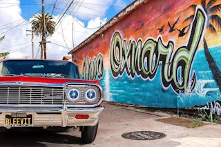 A vibrant red classic car parked by a colorful "Oxnard" mural featuring palm trees, birds, and beautiful ocean scenery.