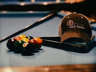 A welcoming pool table with neatly racked billiard balls and a stylish gray Q Club hat, ready for an enjoyable game.