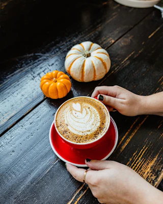 A guest enjoys a beautifully crafted heart latte in a red cup, beside charming pumpkins on a cozy wooden table.
