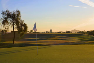Stunning golf course at sunset with a flagstick, rolling green hills, lush trees, and scenic mountains in the background.