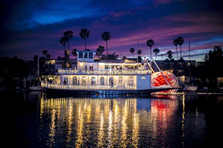 A charming, brightly lit riverboat with a red paddlewheel cruises at dusk, its lights beautifully reflecting on the tranquil water.