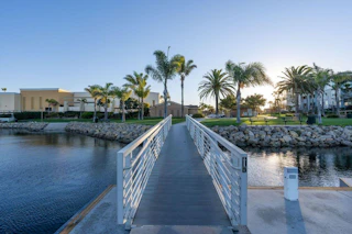 A scenic metal walkway guides visitors past palm trees, lush grass, and charming waterfront buildings as the sun sets beautifully.