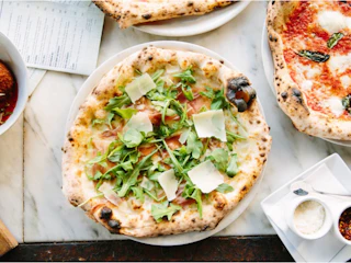 A delicious pizza with fresh arugula, prosciutto, and shaved cheese on a white plate, set among an array of inviting dishes.