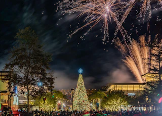 A beautifully decorated Christmas tree glows in a lively plaza as vibrant fireworks illuminate the festive night sky above.