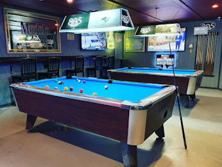 Inviting bar featuring two pool tables, cues, stylish barstools, vibrant wall decor, and TV screens in a lively atmosphere.