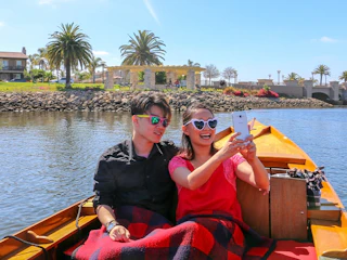 A happy couple enjoys a sunny boat ride, smiling and taking a selfie with lush palm trees in the scenic background.