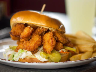 A delicious fried shrimp sandwich with crisp lettuce, pickles, and golden fries served on a white plate, ready to enjoy.