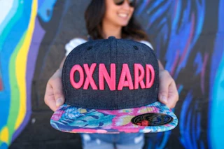 Smiling visitor holding a vibrant "OXNARD" cap with a floral-patterned brim, colorful mural creating a lively backdrop.
