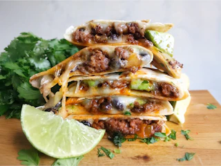 Delicious cheesy beef quesadillas stacked with avocado and black beans, garnished with fresh cilantro and lime on a wooden board.