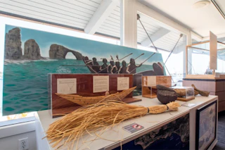 Inviting museum exhibit features a straw boat model, ancient tools, and a vibrant mural of canoeing by scenic coastal rocks.