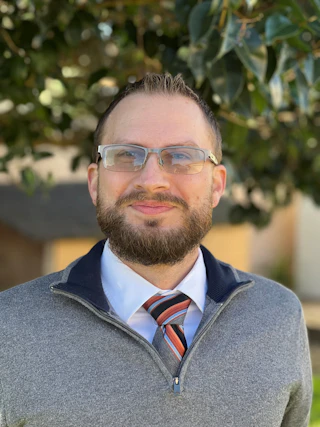 A cheerful man with glasses, a beard, and a striped tie enjoys the outdoors, smiling in front of lush green trees.