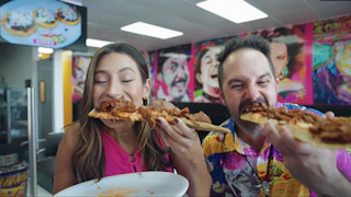 Two people happily enjoy delicious pizza slices in a lively, colorful restaurant decorated with vibrant, artistic wall murals.